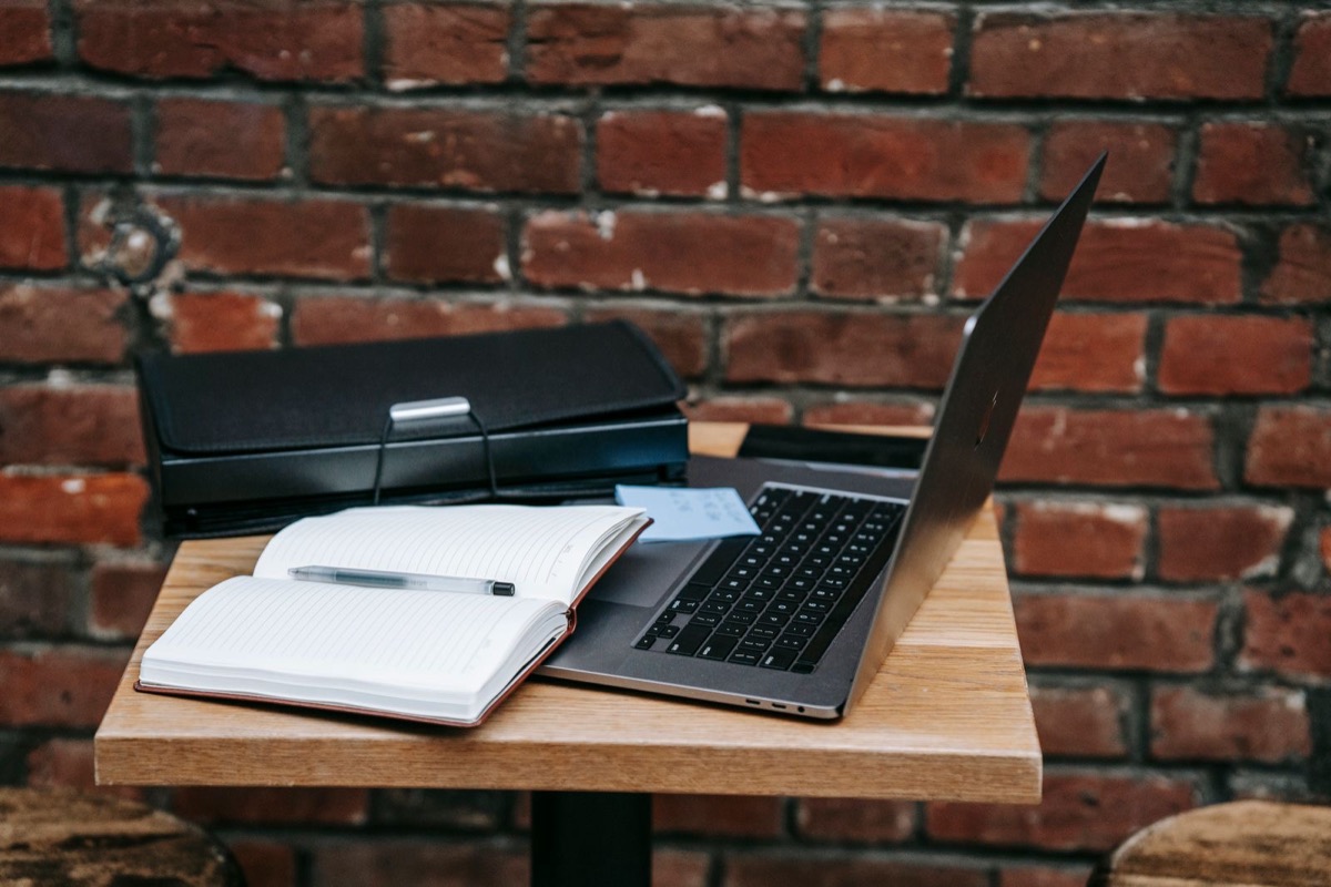 An open notebook and a laptop on a table by a brick wall
